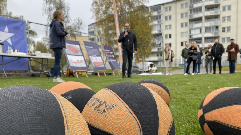Basketbollar på gräsmatta i förgrunden. Hyresbostäders VD Fredrik Edlund i bakgrunden håller invigningstal med mikrofon i handen. Han är svartklädd och står framför byggstängsel med plakat. 
