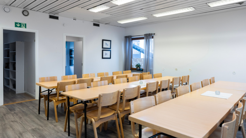 Interiörbild av kvarterslokal Sekreteraren: tre långa träbord med stolar. Ett fönster med grå gardiner och två dörrar som leder till kapprum respektive kök.