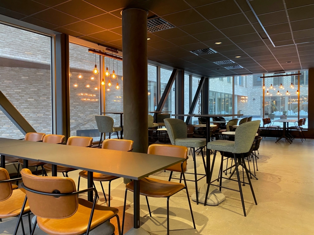 Bild på restaurang inifrån, stora fönsterpartier och bord med tygbeklädda stolar. I fönstret hänger en lampa bestående av flera glödlampor.