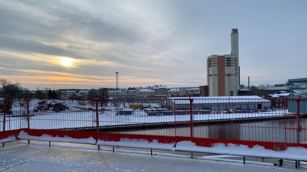 Snöig vy av området Inre hamnen i Norrköping fotat från taket av ett byggprojekt. I förgrunden syns röda byggstaket och vatten (Motala ström). I bakgrunden syns en industribyggnad med hög skorsten.