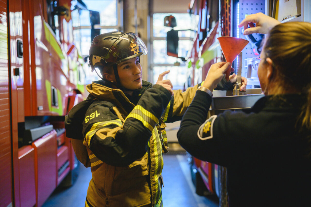 Kvinnlig brandman iförd arbetsklädsel och hjälm står i garaget på en brandstation och pratar med en brunhårig kollega. Brandmannen gestikulerar med armarna i samtalet