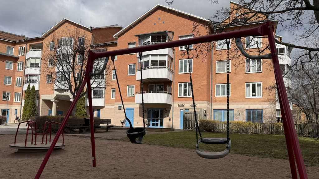 Två gungor på en röd gungställning med ett orangerött lägenhetshus i bakgrunden. 
