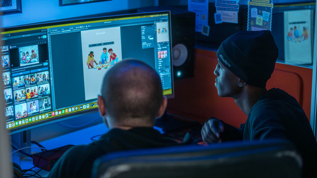 Två män sitter bredvid varandra vid en stor datorskärm i ett nedsläckt arbetsrum. Den ena personen har en mörk mössa på sig.