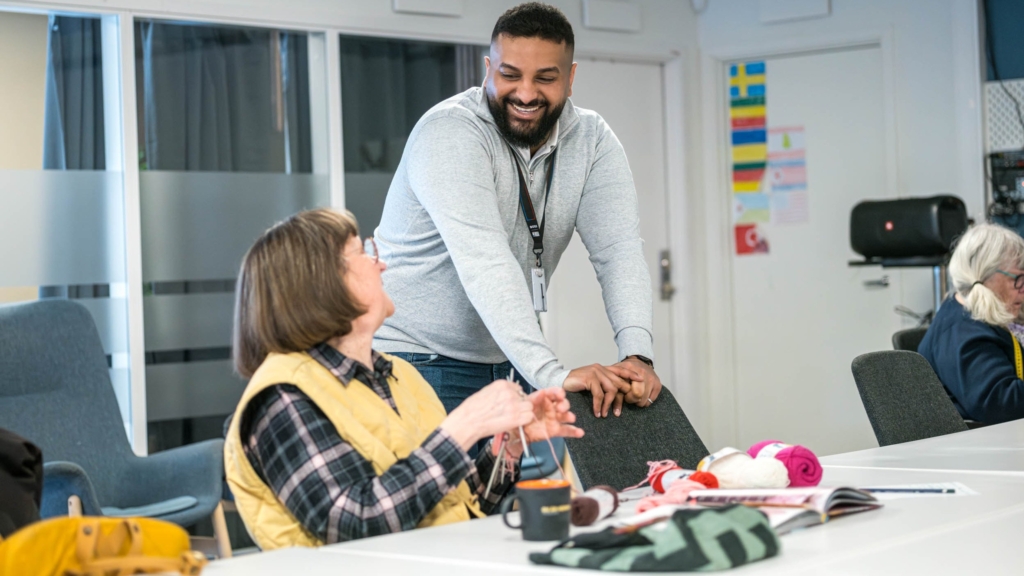 Två personer vid ett bord samtalar. En kvinna sitter på en stol och handarbetar och en man lutar sig mot en stol och ler mot kvinnan.