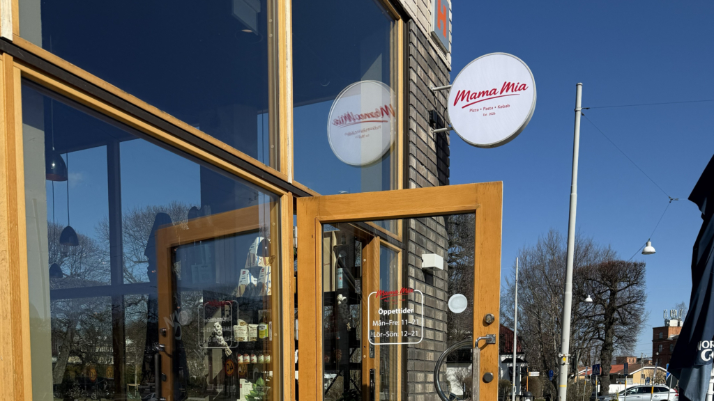 Bord och stolar i trä på uteservering vid restaurang Mama Mia. Blå himmel och rödvitrutig duk på bordet. Dörren till restaurangen på vid gavel