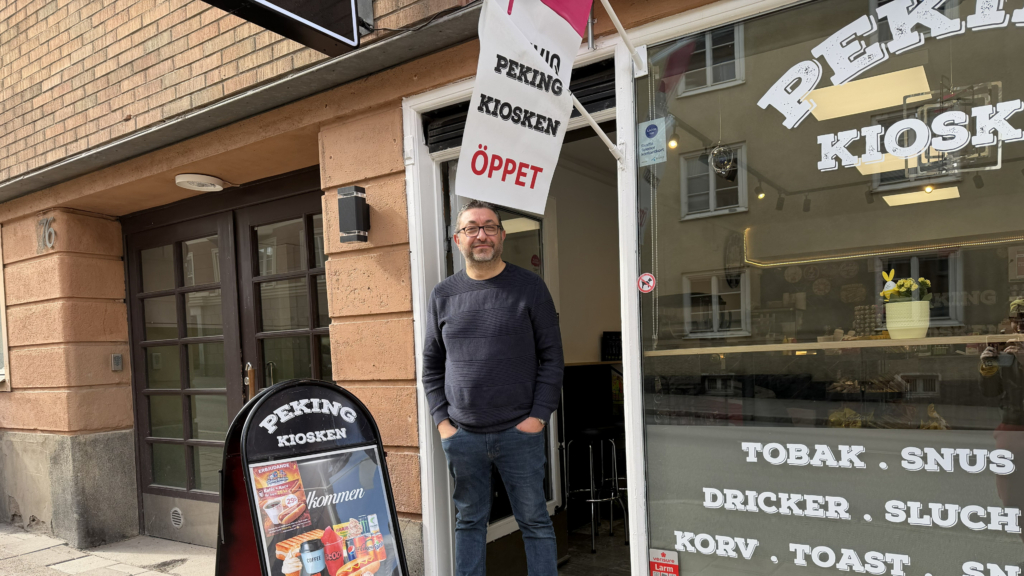 Man i mörkblå tröja och jeans som står med händerna i fickorna i entrén till en kiosk. Kiosken heter Pekingkiosken och har en vägpratare med erbjudanden på gatan utanför och en vit flagga som det står öppet på som sitter på väggen. 