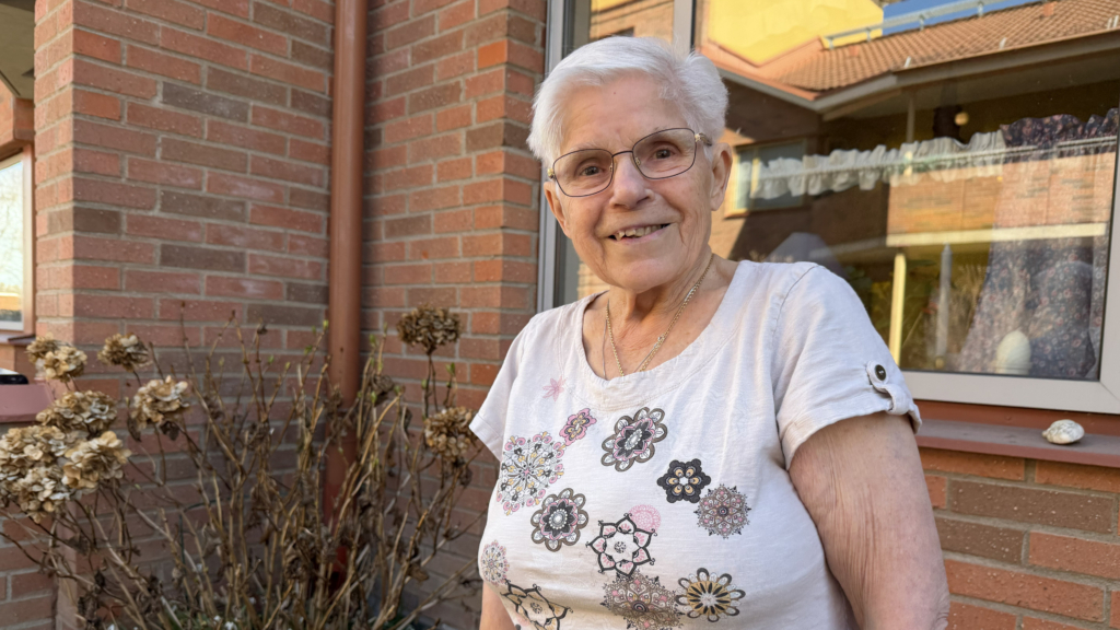 Kvinna med glasögon och vit, blommig t-shirt utanför byggnad i rött tegel och stora fönster. Tittar in i kameran och ler