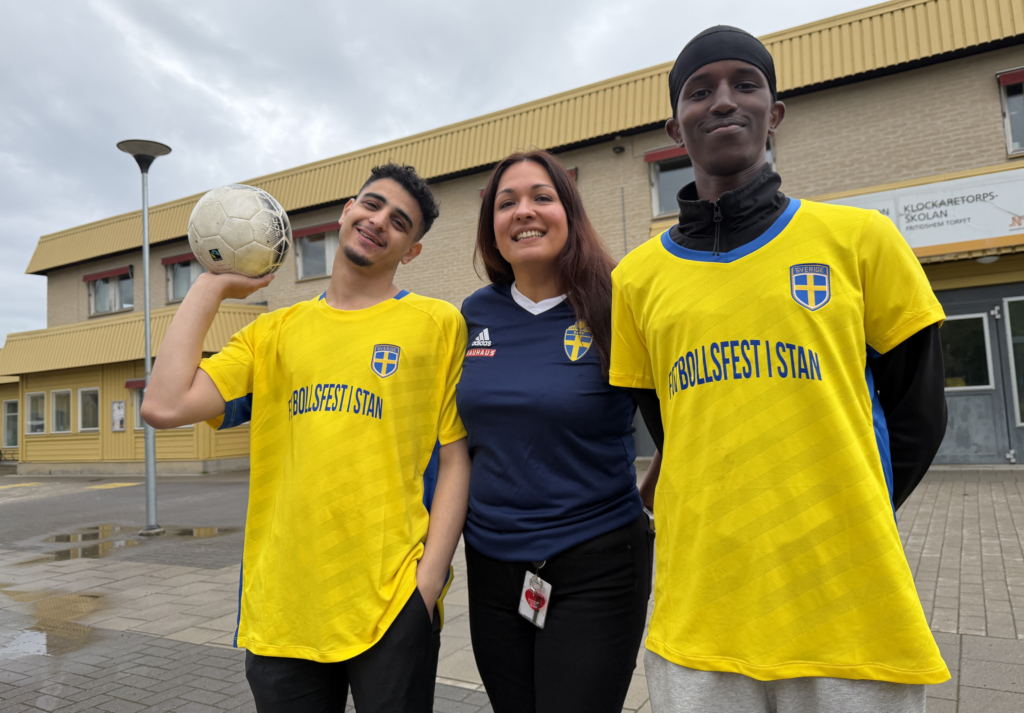 Tre personer med fotbollströjor poserar glatt framför kameran med ett gult hus i bakgrunden. Från vänster: man med gul Sverige-fotbollströja, kort brunt hår och fotboll i handen, i mitten kvinna med blå Sverigetröja och brunt, långt hår, till höger man med gul Sverigetröja och svart huvudbonad.