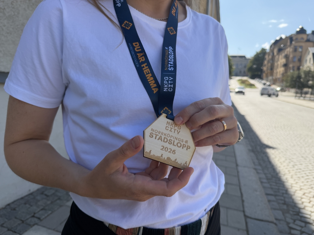 Kvinna i vit t-shirt håller i en medalj som hänger runt halsen i ett blått band. Medaljen är gjord i trä och har testen stadsloppet 2026 på.