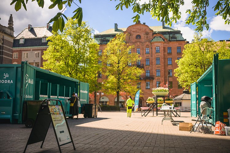 Torgyta med containers uppställda, innehållandes sopkärl. I bakgrunden ett rött tegelhus och i förgrunden en gatupratare. Vårgröna träd utsmyckar parken.