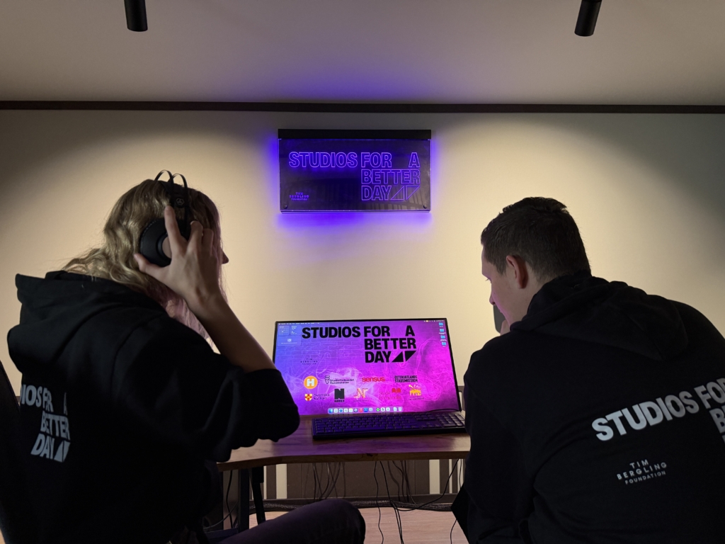 Två människor med svarta hoodies med texten Studios for a better day på ryggen sitter i en nedsläckt studiomiljö och lyssnar på musik
