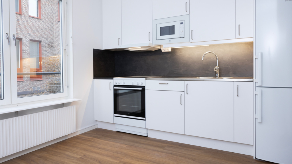 Interiörbild av nyrenoverad lägenhet på studentboende Dalkarlen. Ett kök med vita luckor, kyl och frys samt ett stort fönster på vänster sida av köket.
