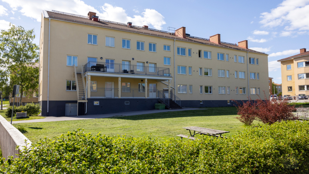 Exteriörbild av studentboendet Assessorn i stasdelen Klingsberg. I förgrunden är utemiljön med buskar, gräsmatta och bänkar.